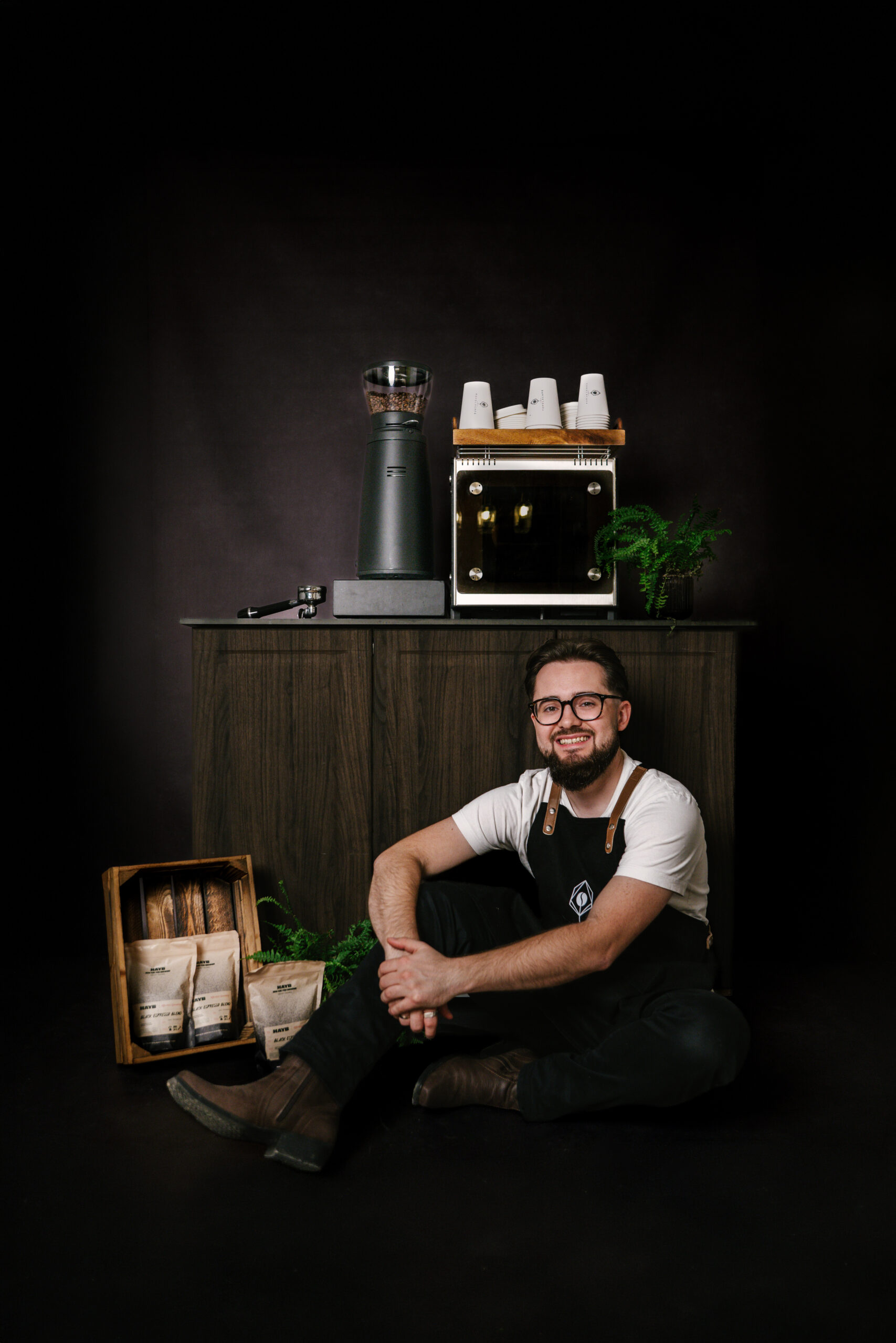Topola Cafe barista na tle wózka kawowego siedzi na ziemi, w tle kawa i ekspres dalla corte min, obok ekspresu młynek Compak i3 pro